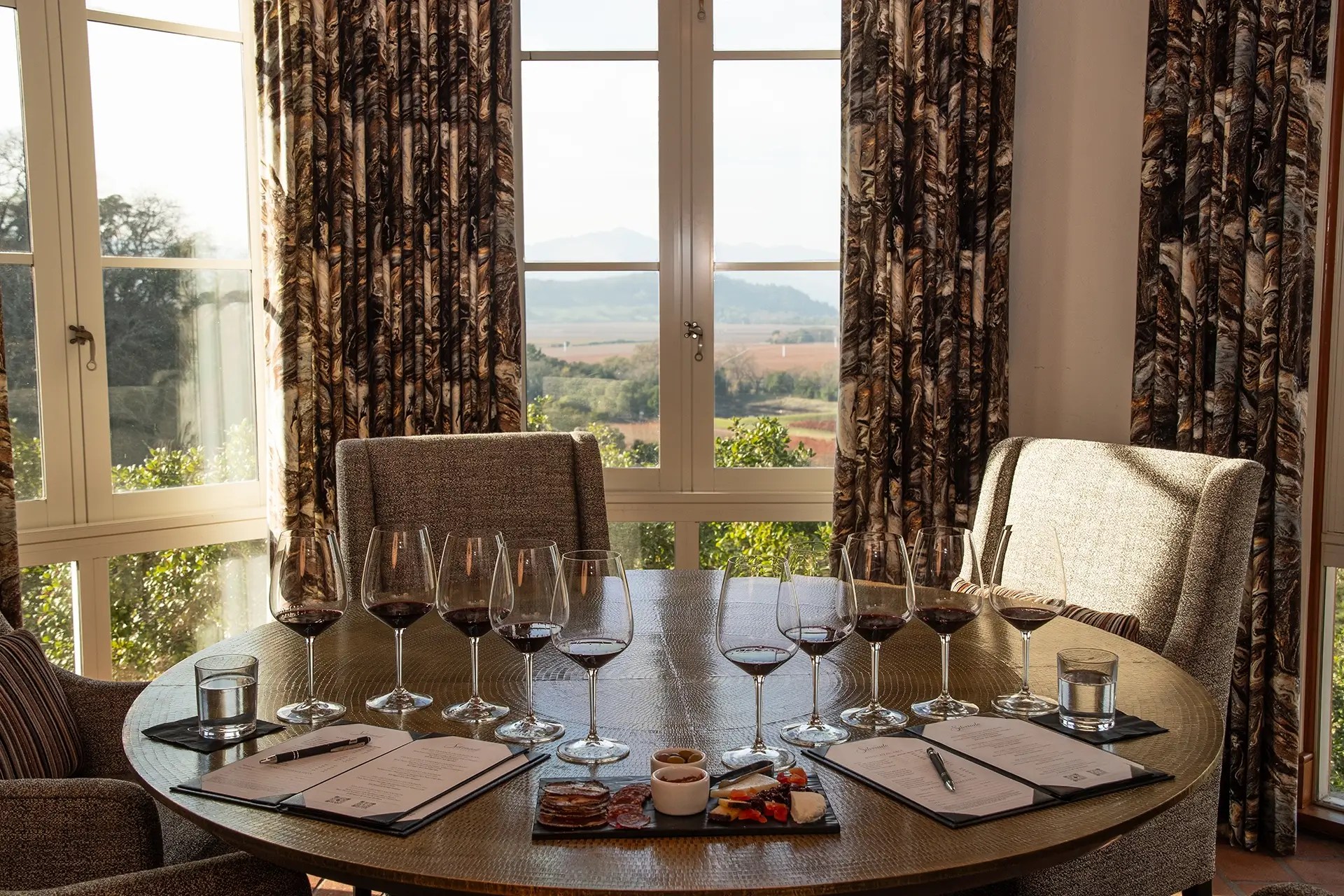 Wine tasting table set with glasses and charcuterie beside vineyard-view windows